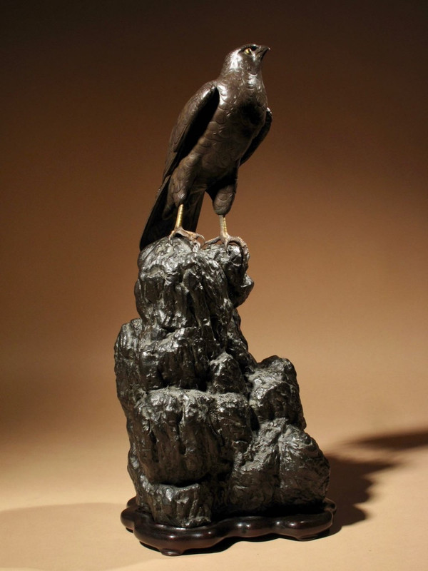 A highly decorative bronze model of a hawk sitting on a steep rock.